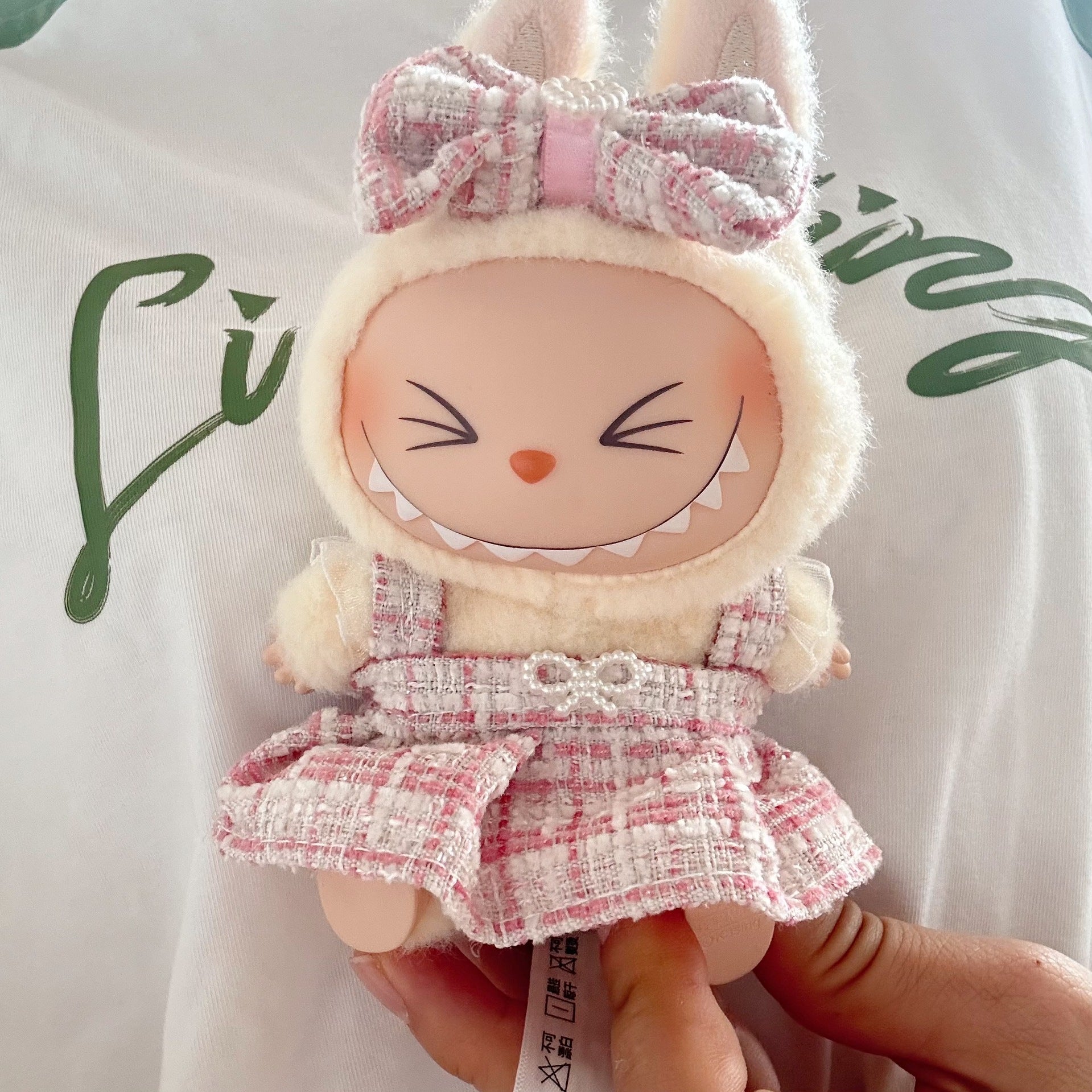 Plush toy with bunny ears and a pink dress held by a hand on a white background with green text.