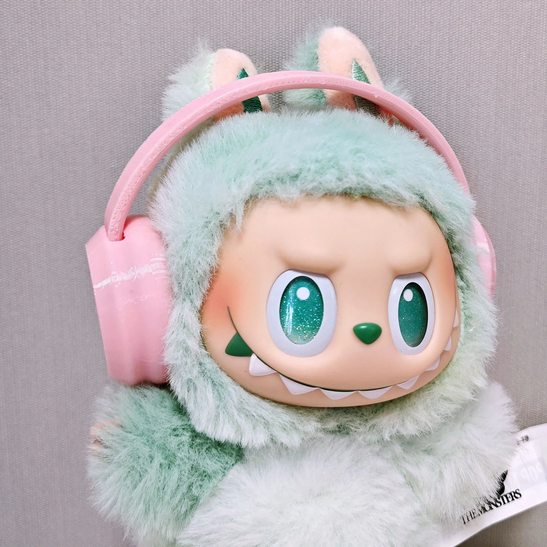 Plush toy with green fur and pink headphones on a gray background
