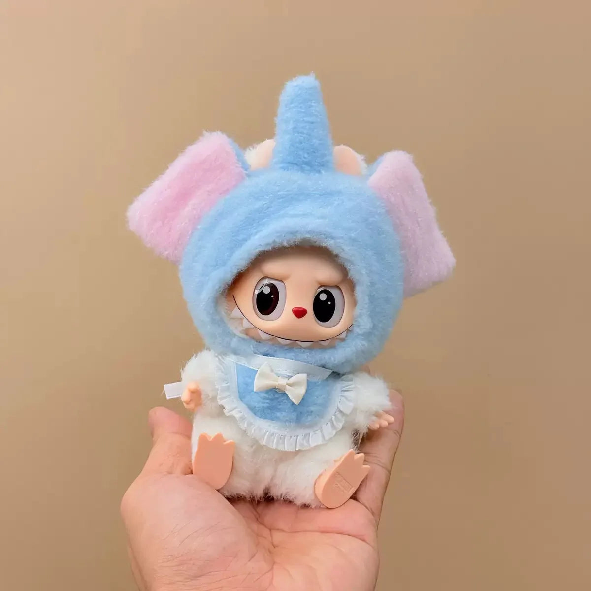 Small plush toy with a blue hood and pink ears held in a hand against a beige background