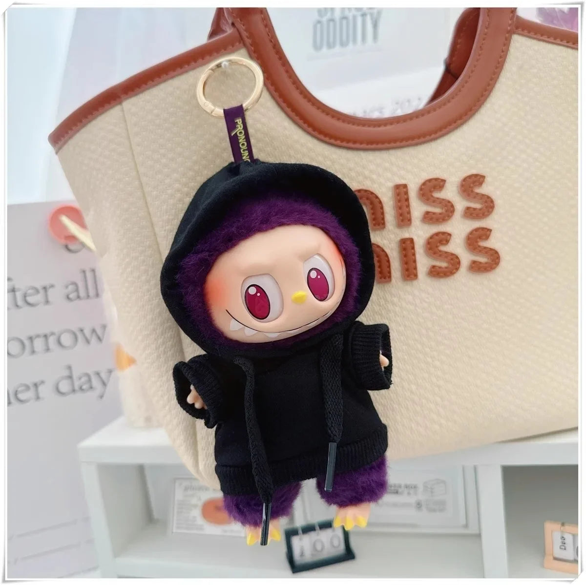 Plush keychain with a purple hood and black outfit in front of a beige bag with 'MISS' text.