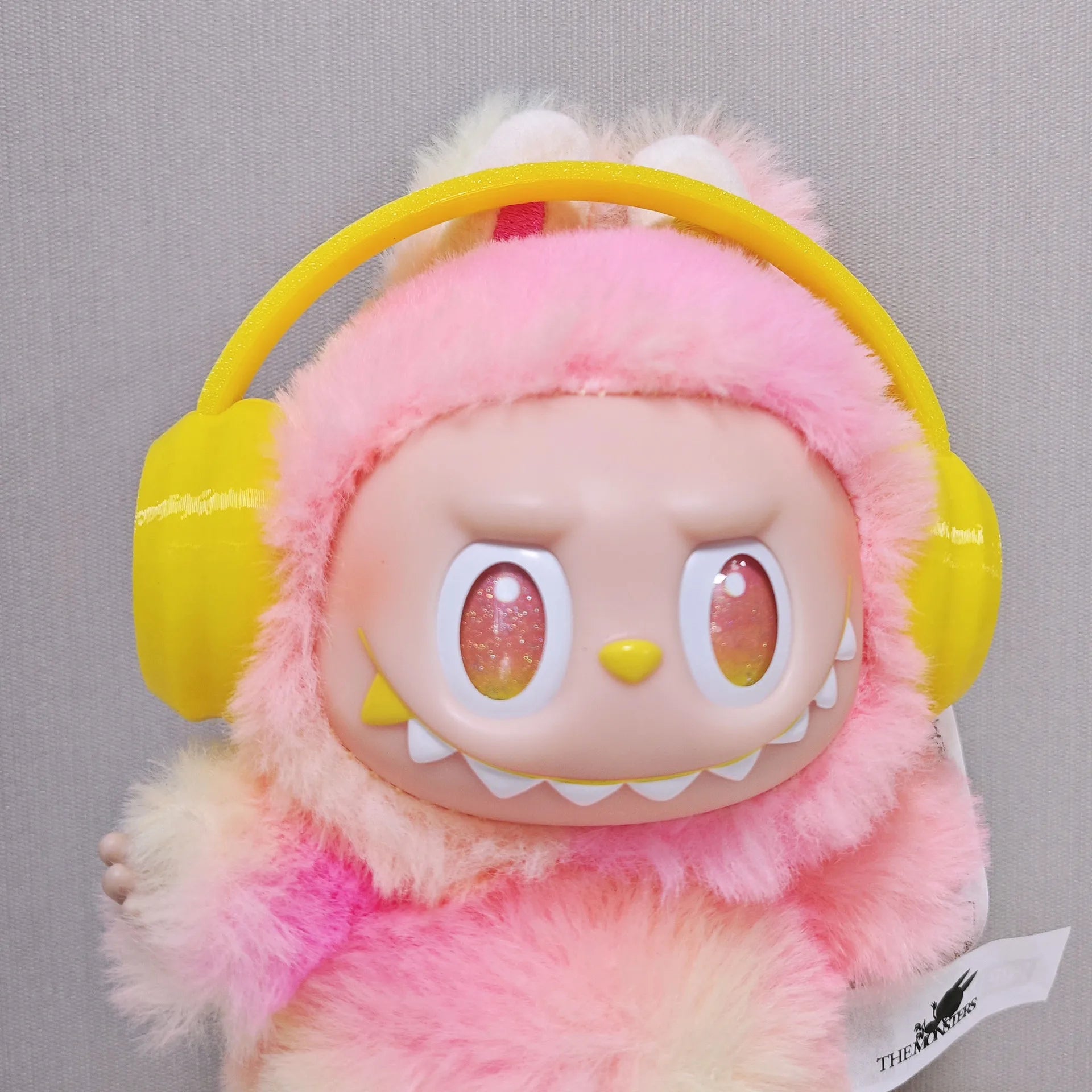 Plush toy with pink fur, yellow headphones, and a tag on a gray background
