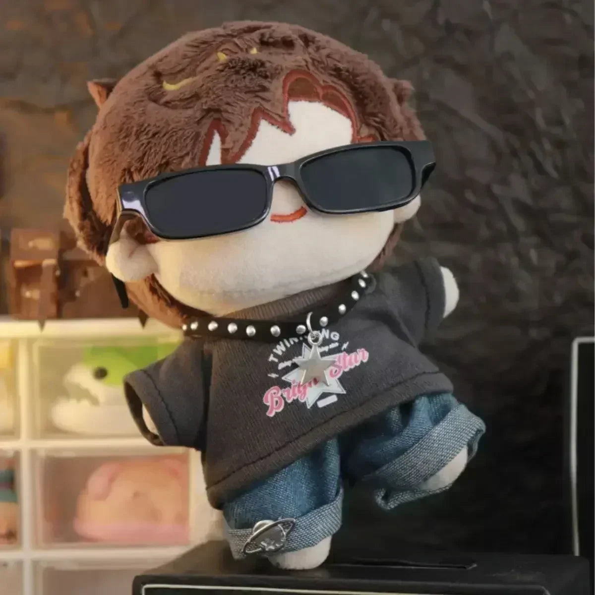 Plush toy with sunglasses and a black shirt with pink text, standing on a surface with a blurred background.