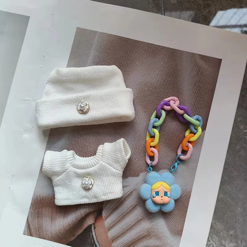 White baby outfit with hat and sweater, colorful teething toy, and blue flower-shaped pacifier clip on a textured surface.