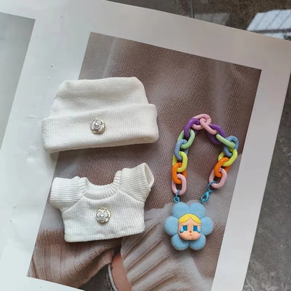 White baby outfit with hat and sweater, colorful teething toy, and blue flower-shaped pacifier clip on a textured surface.