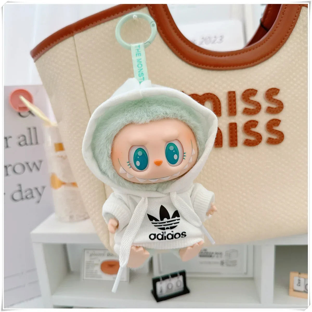 Plush toy with a hoodie and Adidas logo in front of a beige bag with 'MISS' text.
